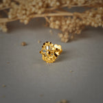 A gold cutwork nose pin with a floral open design and a beaded lower edge, placed on a grey background with dried flowers