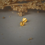 A gold flower-shaped stud earring with rounded petals and a beaded center, placed on a grey background with dried flowers