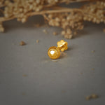 A round gold nose pin with a ribbed edge and a faceted center, placed on a grey background with dried flowers