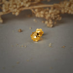A gold flower-shaped stud earring with a textured petal design, placed on a grey background with dried flowers