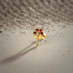 An 18K gold nose pin shaped like a flower with ruby-red petals and a clear crystal center, with two hanging clear crystal drops below, photographed on a light textured surface