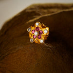 An 18K gold nose pin shaped like a dense flower cluster with small heart-shaped gold accents, set with alternating ruby-red and clear crystal stones, photographed on a dark textured fabric background