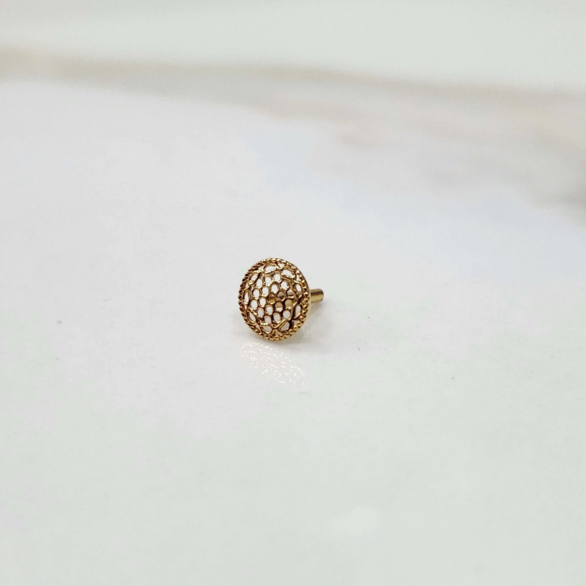 Front view of a small round 18K nose pin gold placed on a white surface highlighting its detailed texture

