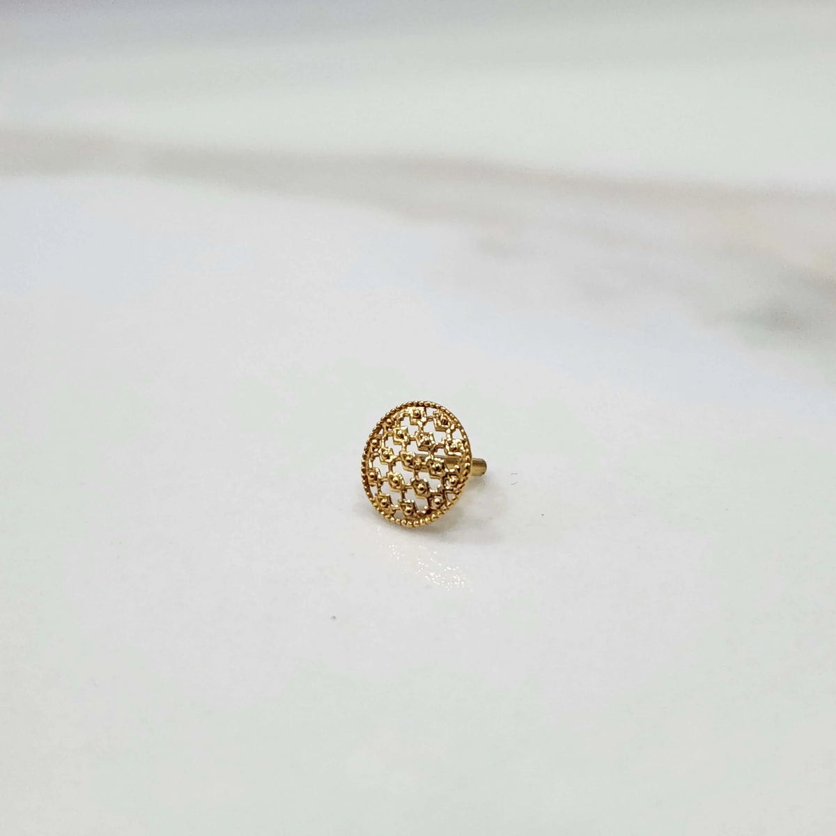 Front view of small textured 18K gold nose pin placed on a white surface highlighting its fine detailing