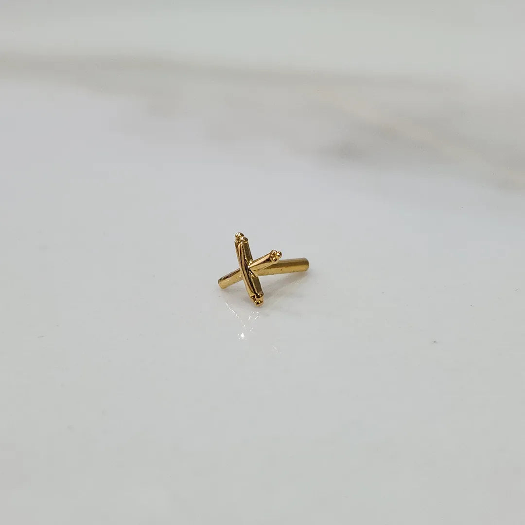 Angled close up of a small cross shaped gold nose pin on a white background highlighting fine detailing