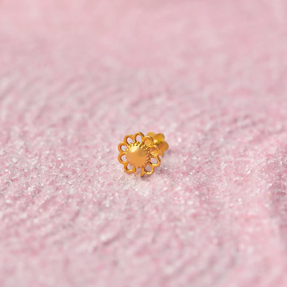 Close up of a floral inspired 18K gold nose pin placed on a soft pink textured background highlighting its intricate design