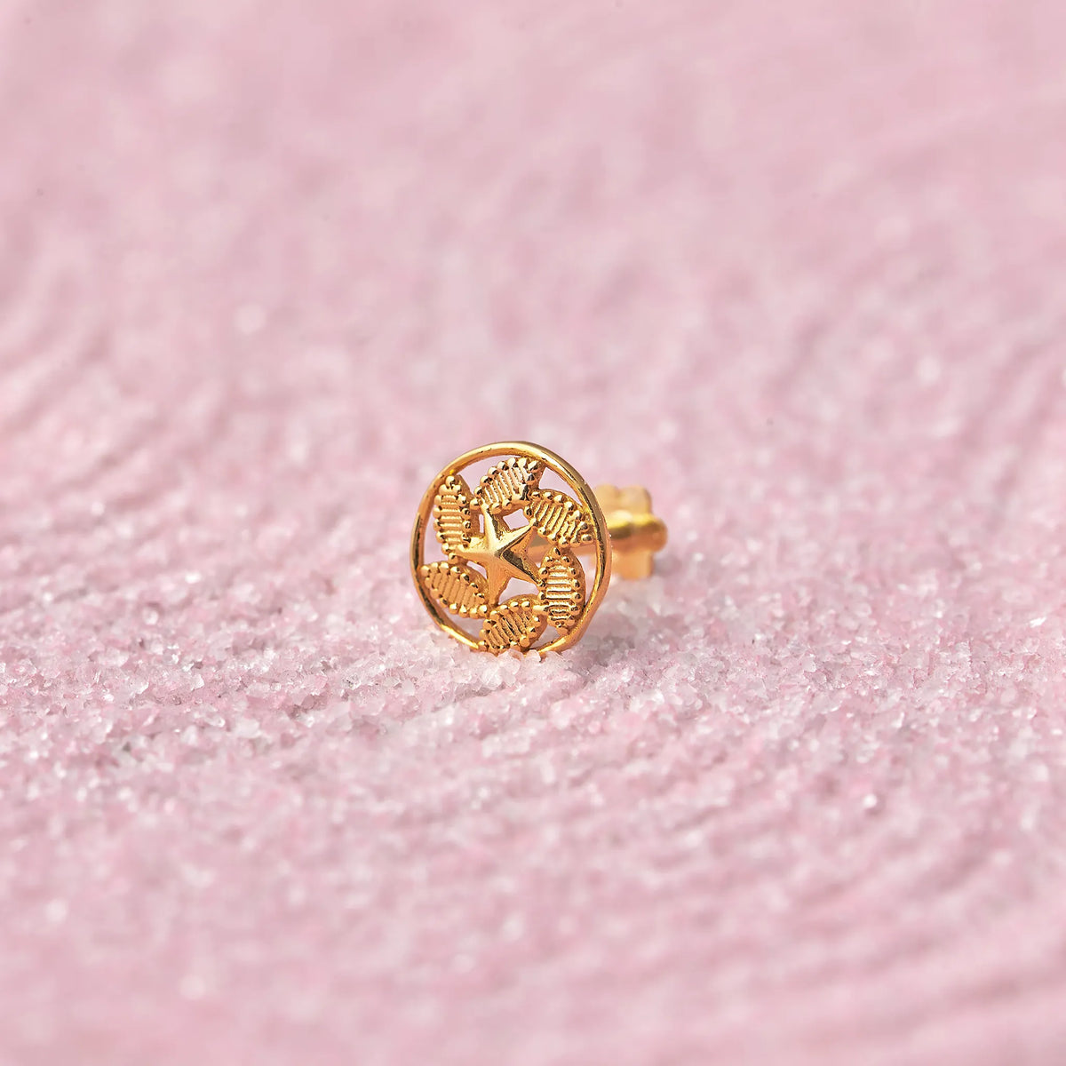 Close up of a round patterned 18K gold nose pin placed on a soft pink textured background highlighting detailed craftsmanship