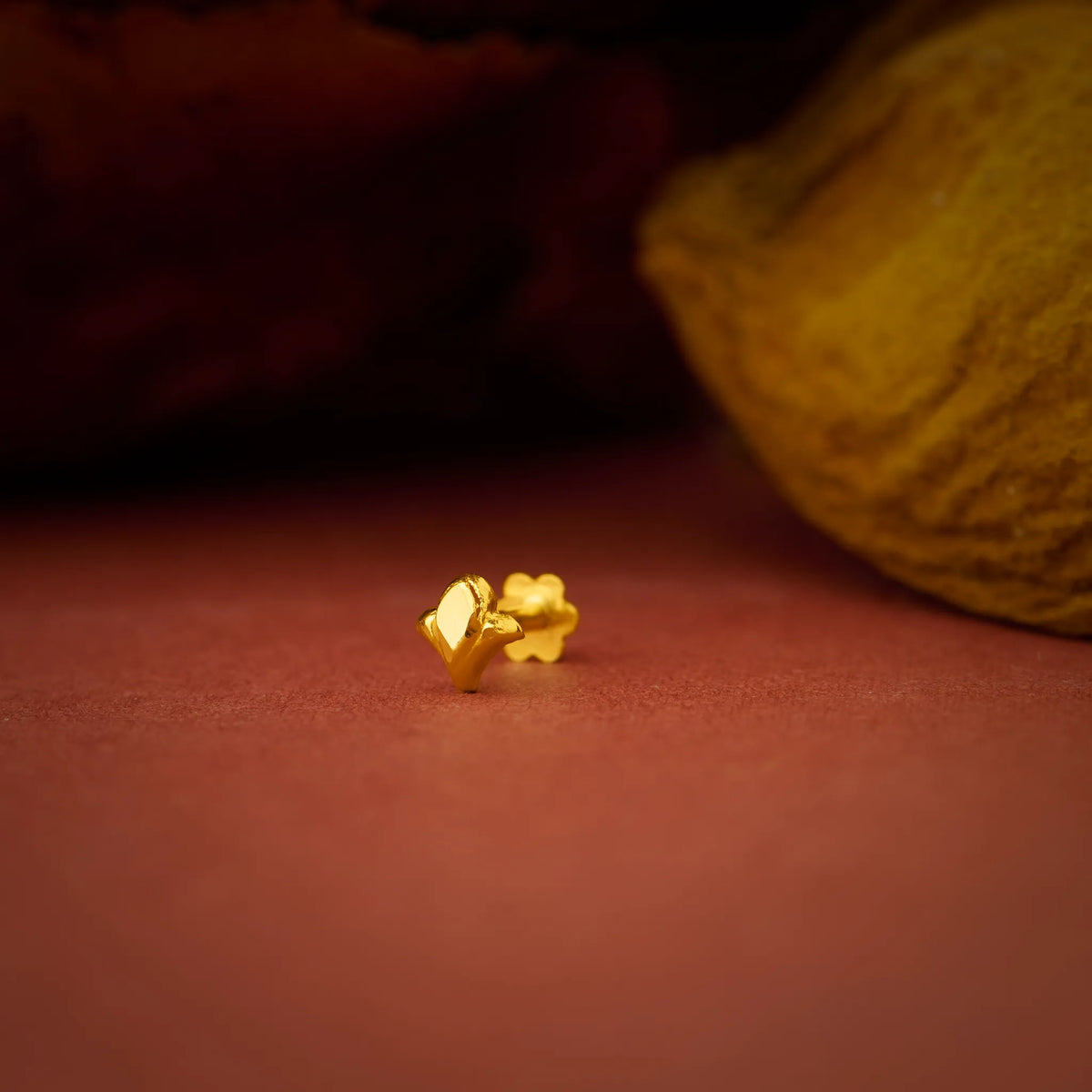Close up of small leaf inspired 18K gold nose pin placed on a warm terracotta surface highlighting its smooth polished finish