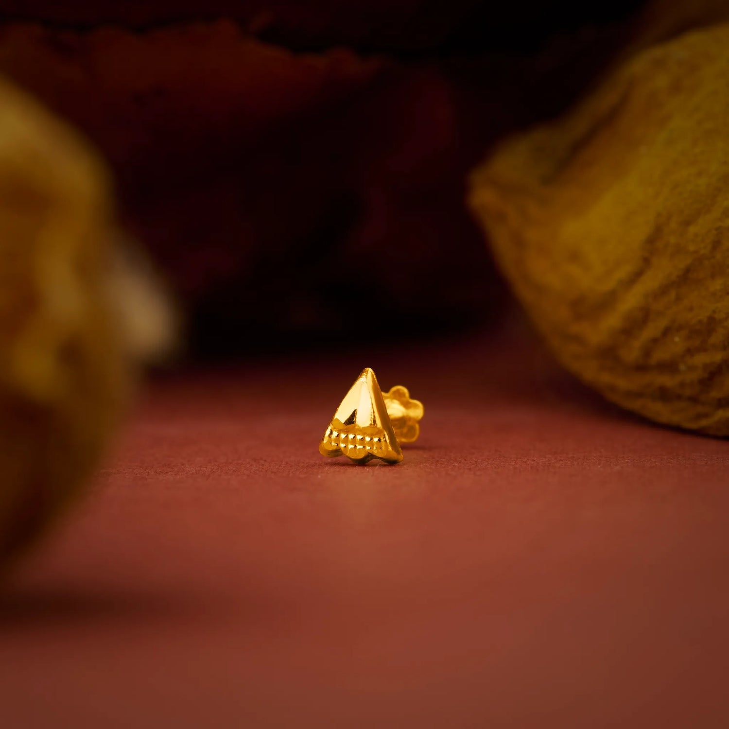 Close up of a tiny triangular gold nose pin placed on a warm brown surface highlighting its sharp geometric shape and polished finish