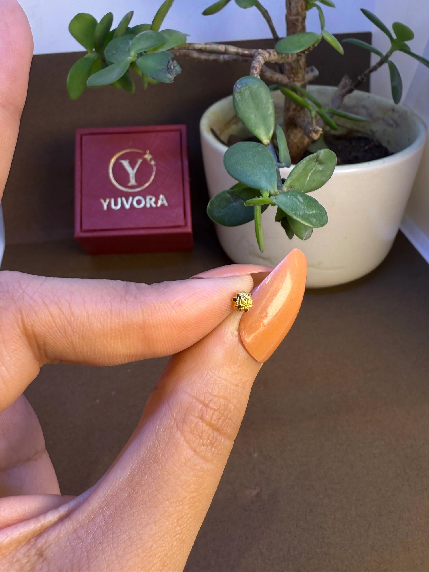 Hand holding a tiny gold nose pin near a Yuvora box showing its lightweight feel and wearable size