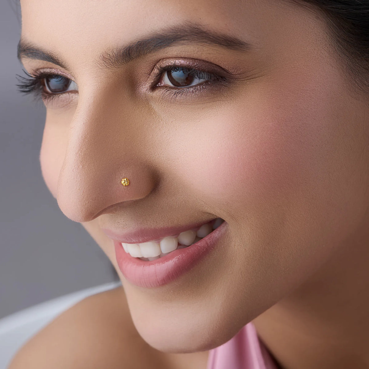 Close-up of a woman's face with a gold minimal nose pin or nose ring made with 18 karats real gold against a neutral background 