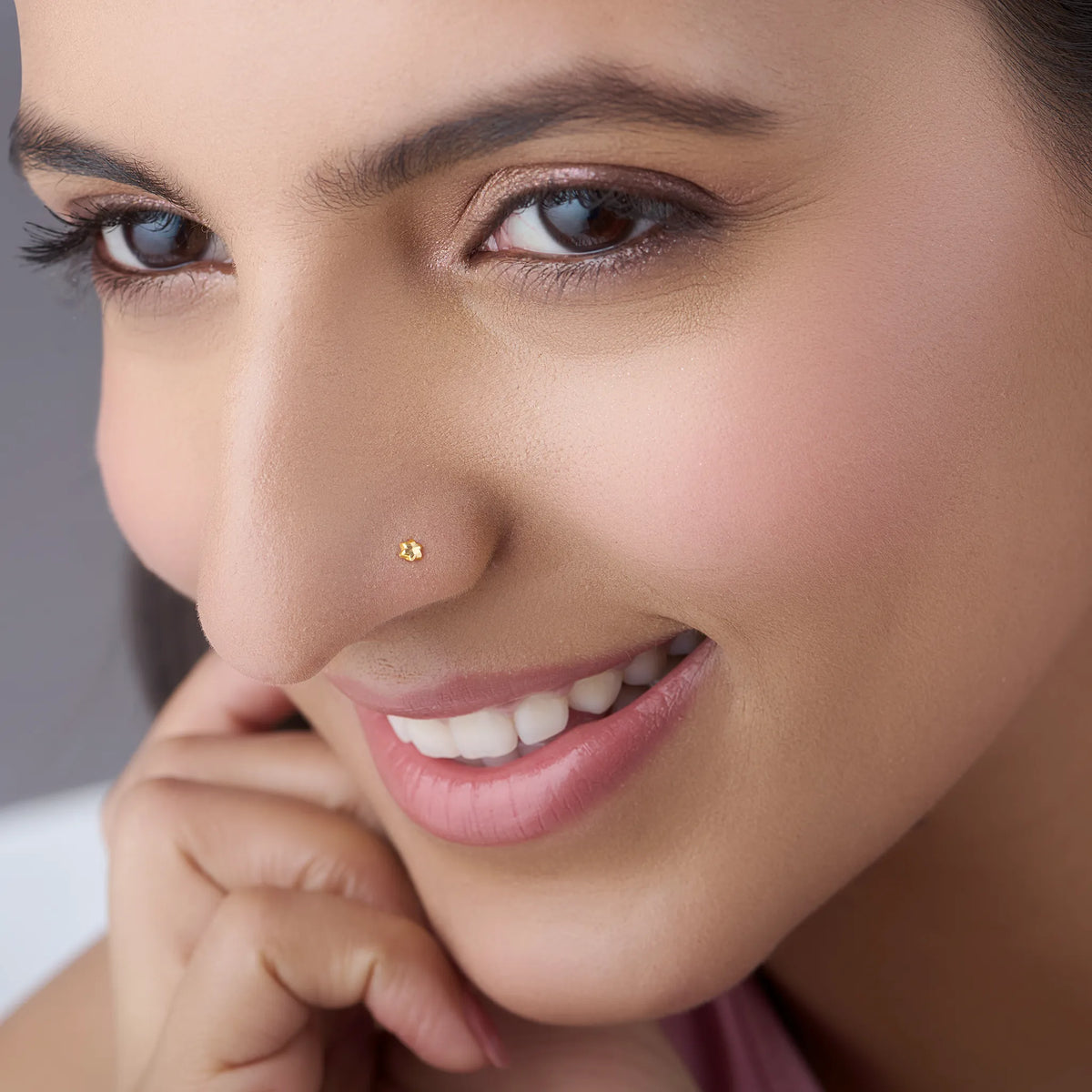 Smiling woman wearing a delicate 18K gold nose pin shown in close up emphasizing subtle shine and comfort fit