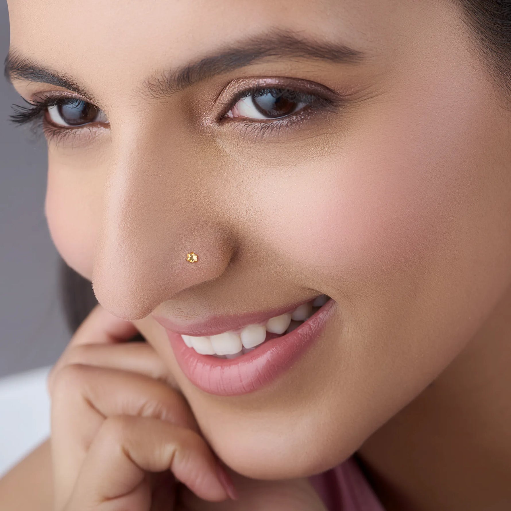 Smiling woman wearing a delicate 18K gold nose pin shown in close up emphasizing subtle shine and comfort fit