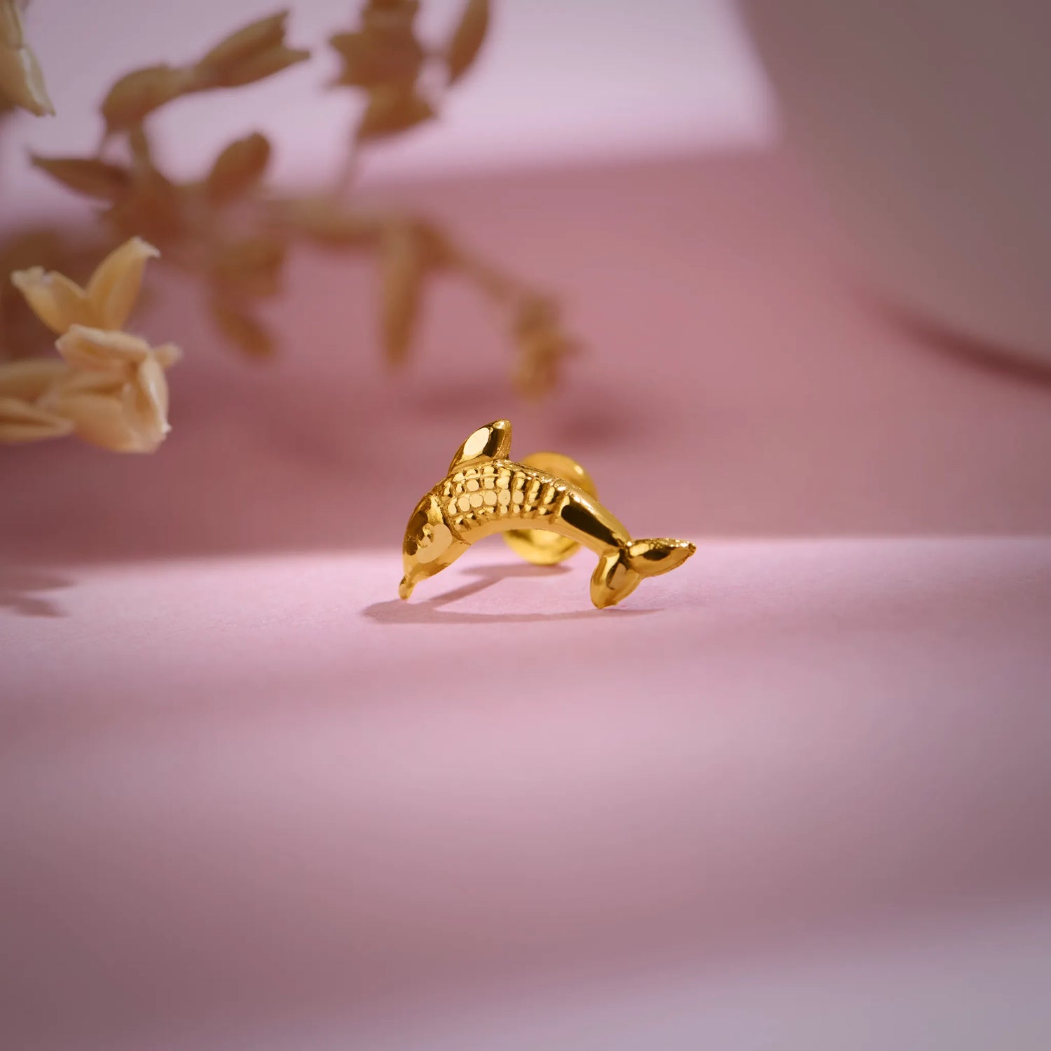Dolphin-shaped 18K nose pin gold photographed on a soft pink background showing its fine detailing
