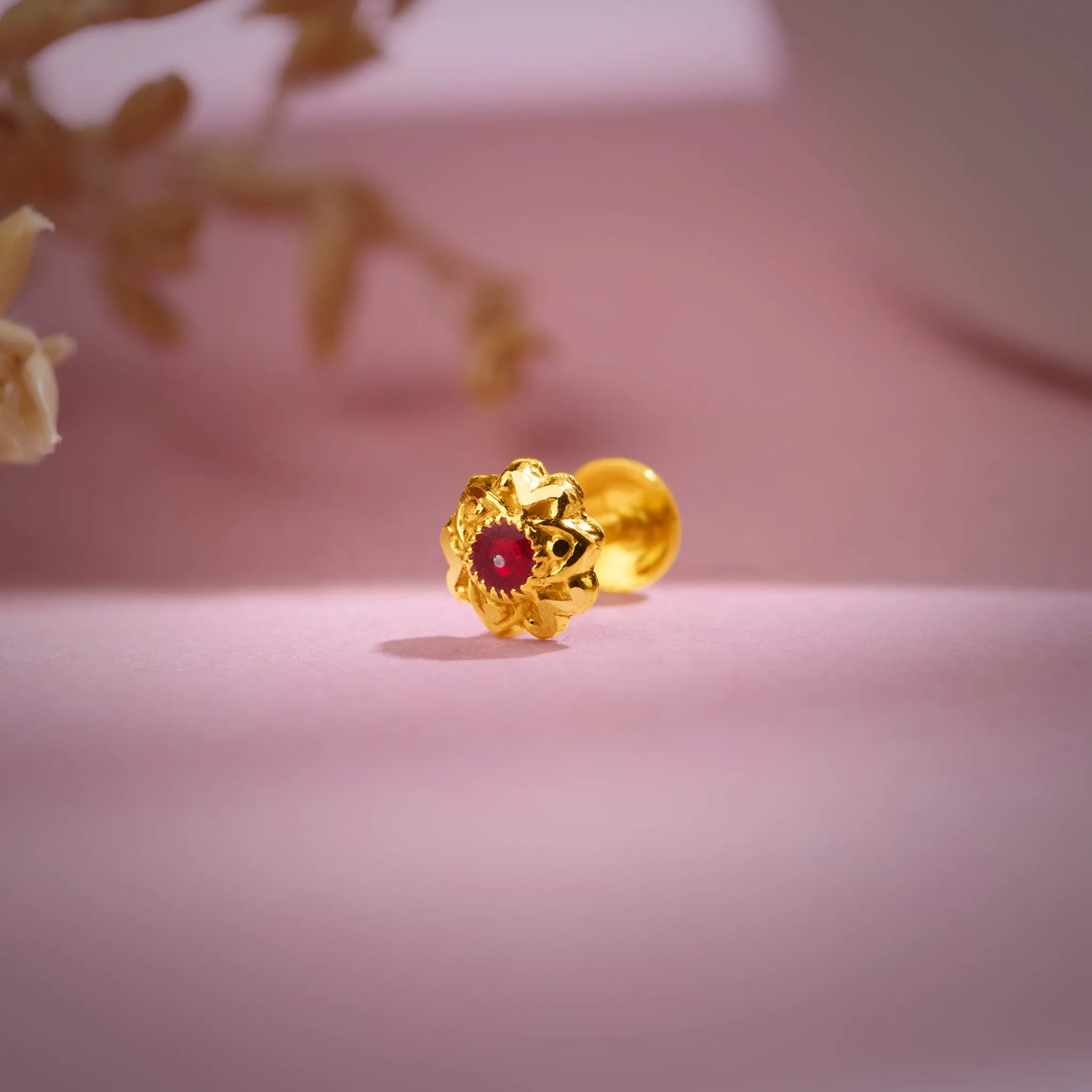 Gold nose pin with a red stone placed on a soft pink surface showcasing detailed floral design