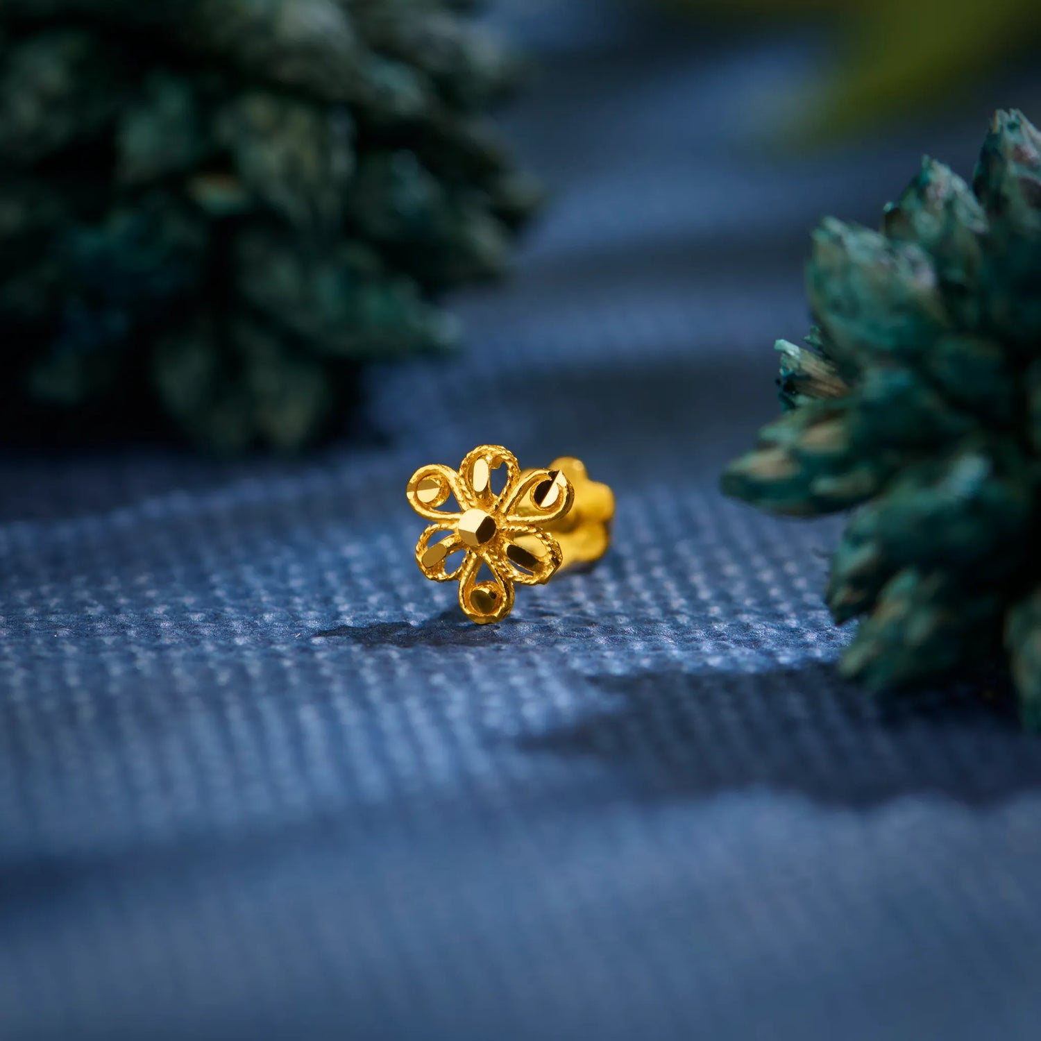 Close up of a floral 18K nose pin gold placed on a textured blue surface with greenery highlighting intricate handcrafted detailing