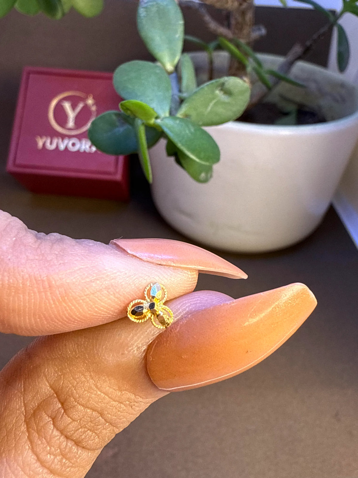 Close up of a hand holding a small multi stone floral 18K gold nose pin between fingers with Yuvora branding and a potted plant in the backgroundv