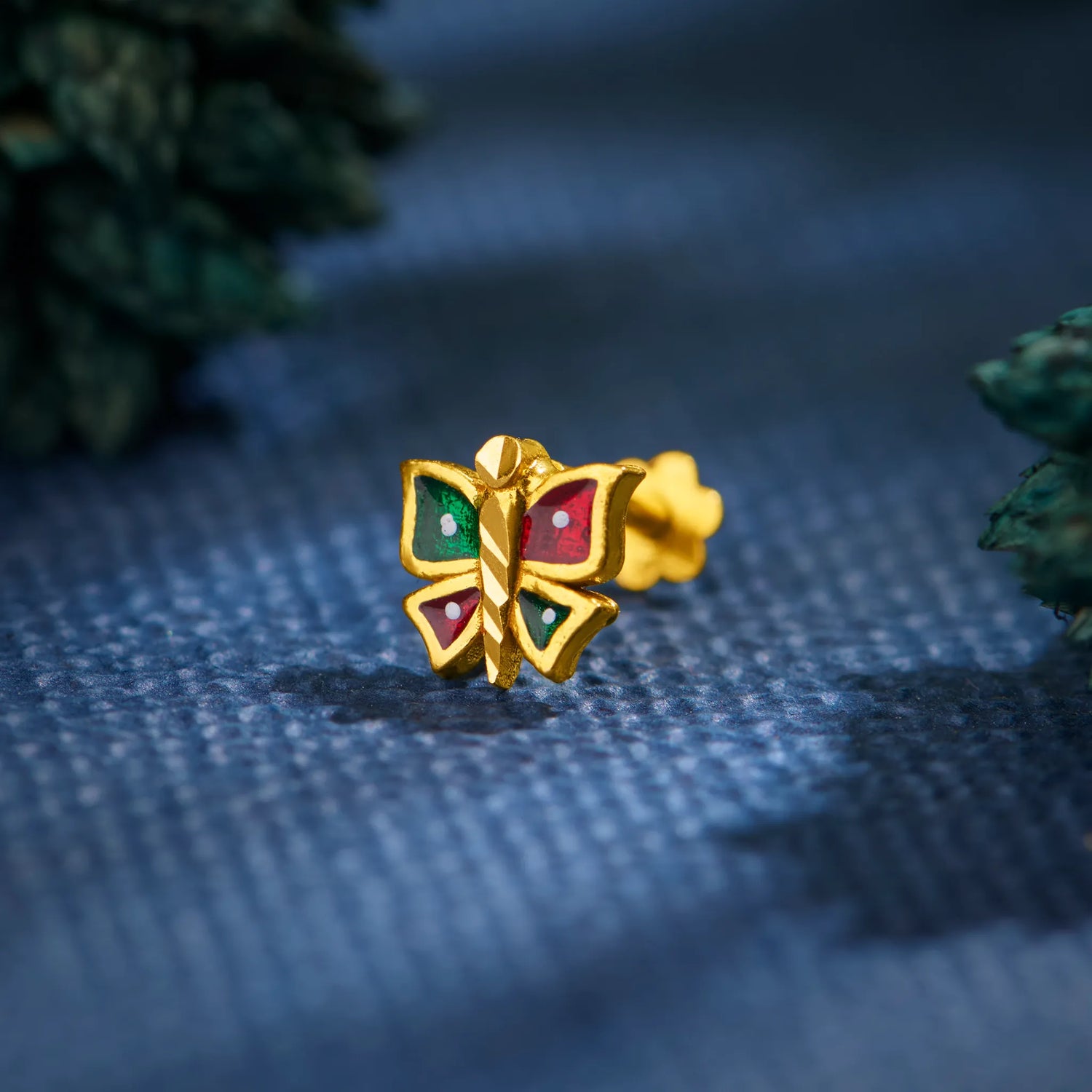 Close-up of multicolour butterfly-inspired 18K gold nose pin placed on a textured blue surface highlighting its vibrant enamel detailing