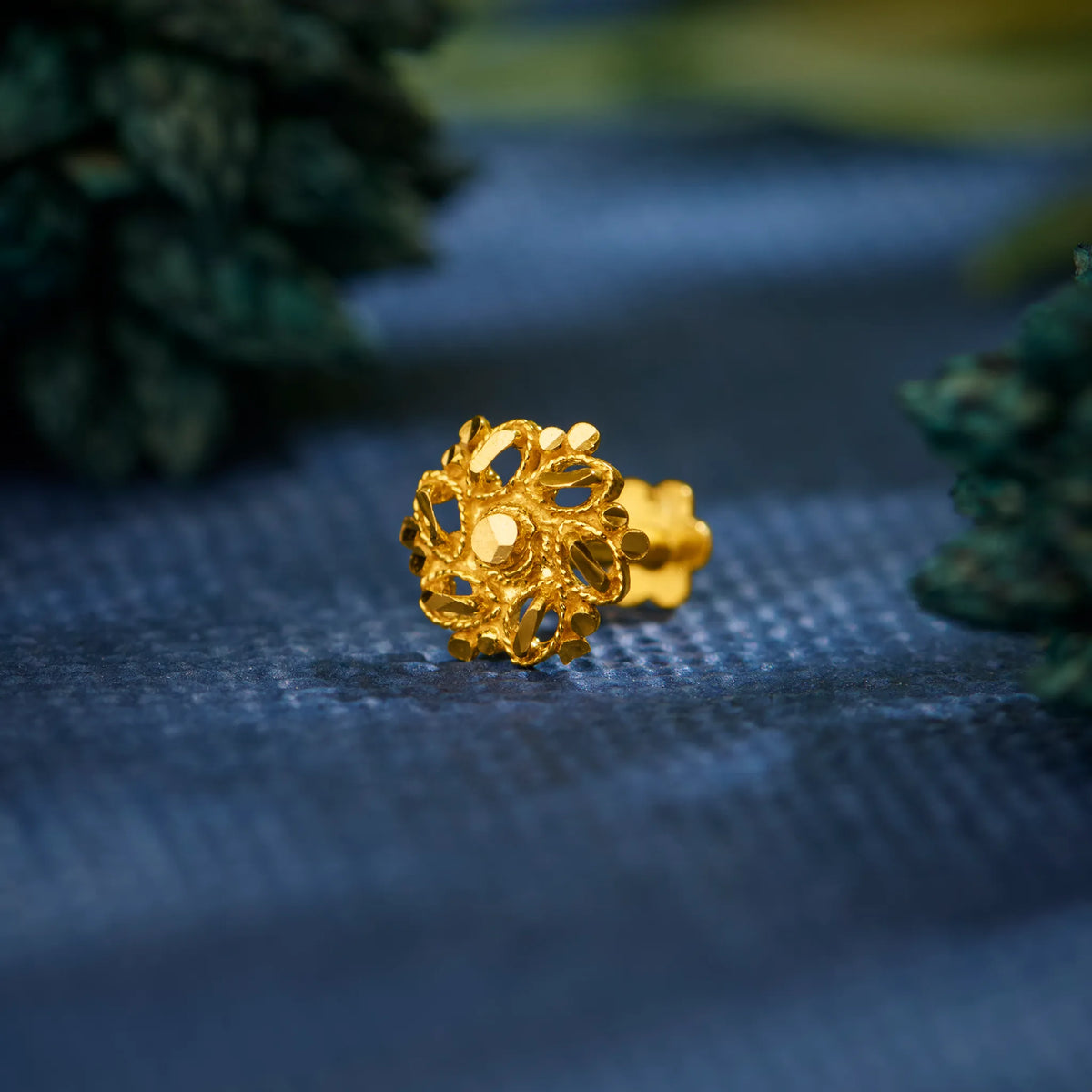 Close up of an intricate 18K nose pin gold with floral design placed on a textured blue surface highlighting fine craftsmanship