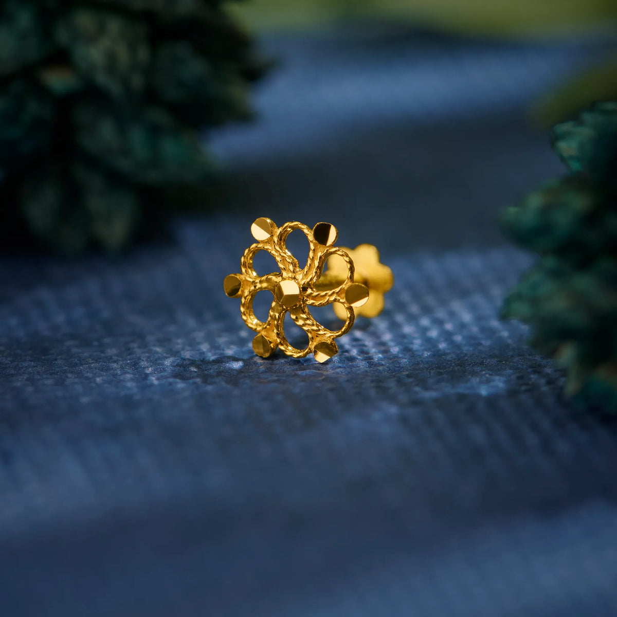 Close up of a floral 18K nose pin gold placed on a textured blue surface highlighting its intricate openwork design