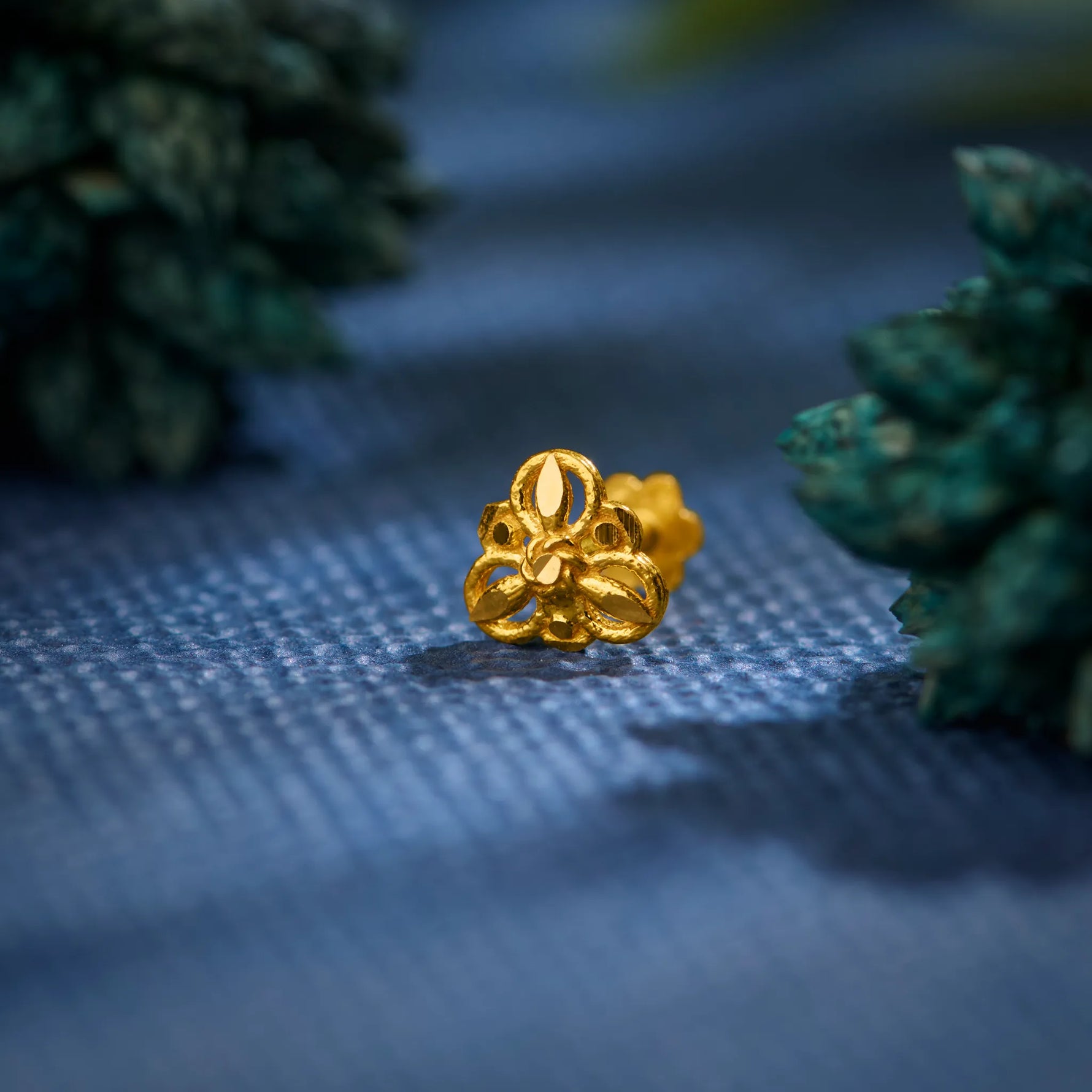Close-up of a handcrafted 18K gold nose pin placed on a textured blue surface surrounded by soft greenery