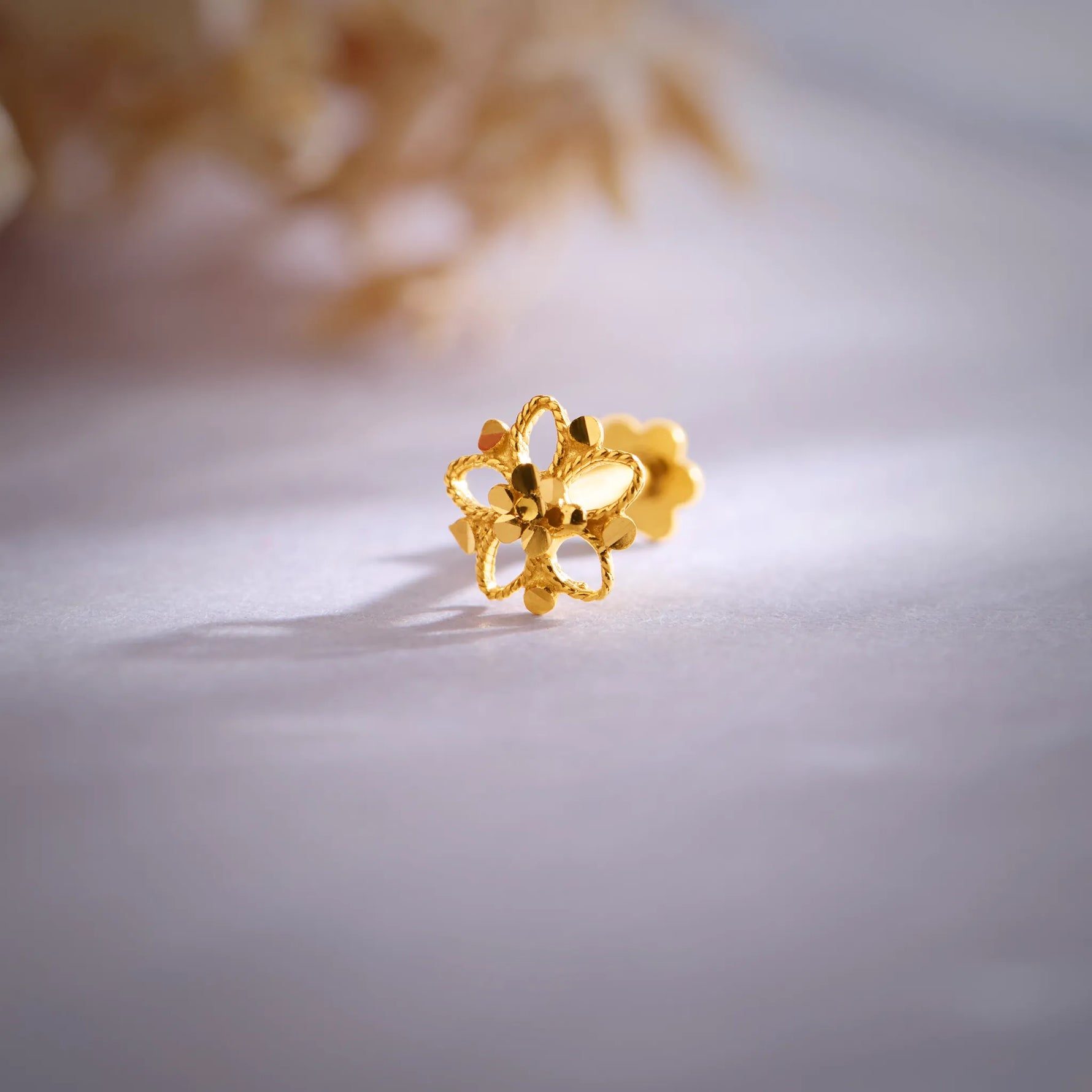 Close up of floral patterned 18K gold nose pin placed on a soft lavender surface highlighting its intricate detailing