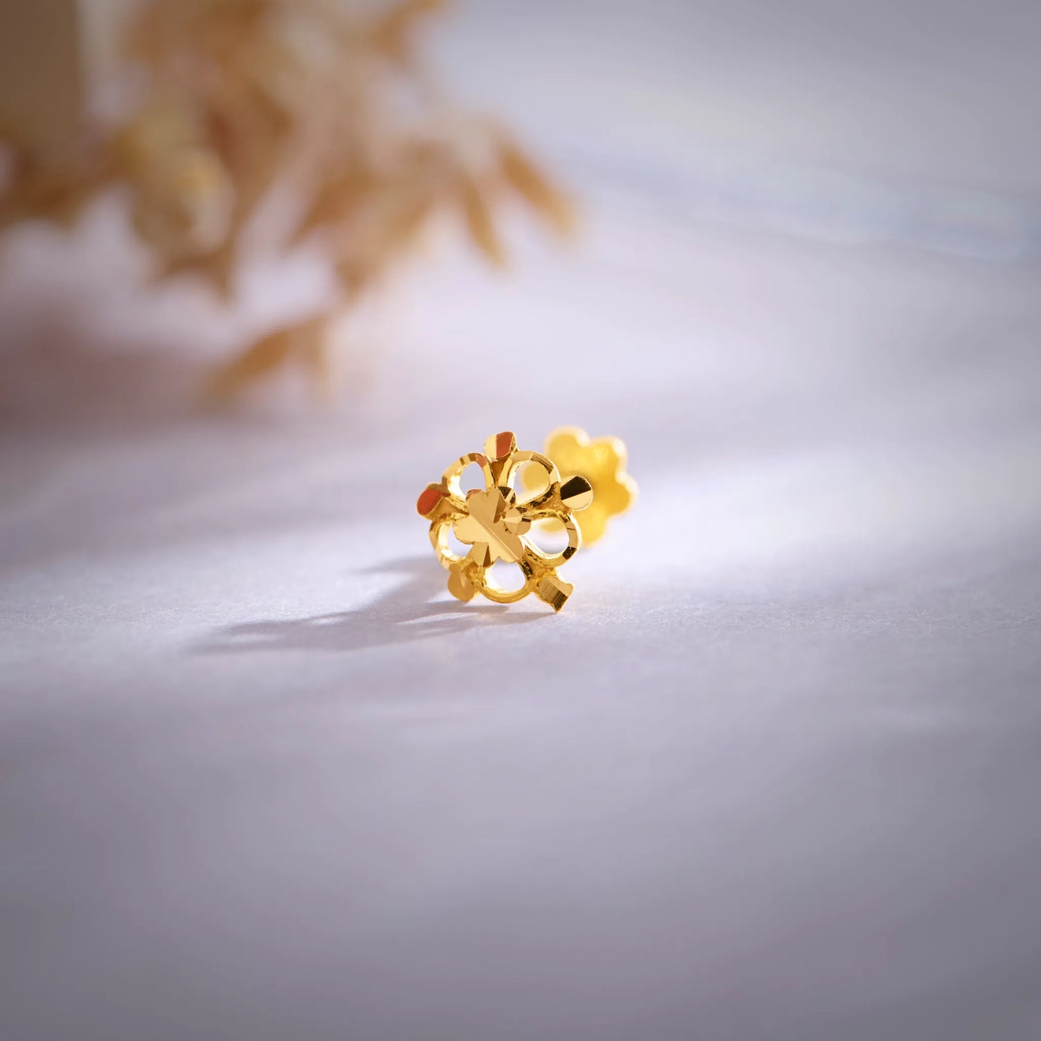 Close-up view of an 18K gold floral nose pin placed on a soft pastel surface showcasing its intricate petal design