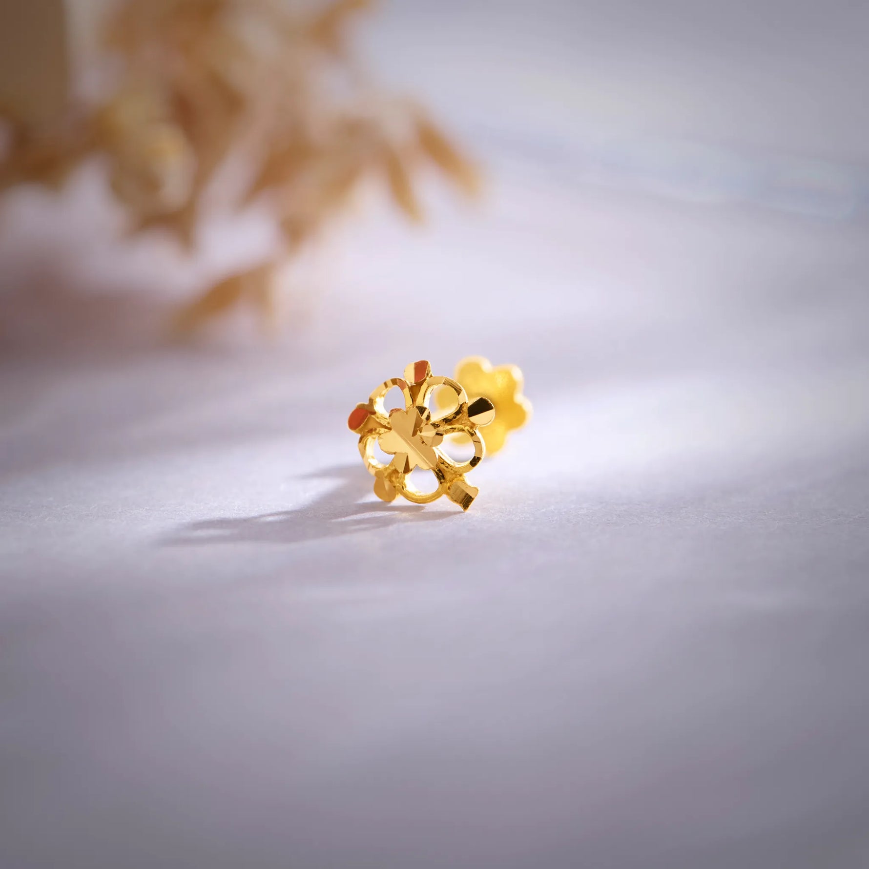 Close-up view of an 18K gold floral nose pin placed on a soft pastel surface showcasing its intricate petal design
