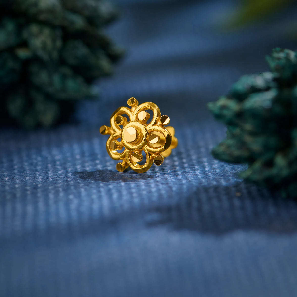 Close up of floral patterned 18K gold nose pin placed on a blue textured surface highlighting its intricate detailing