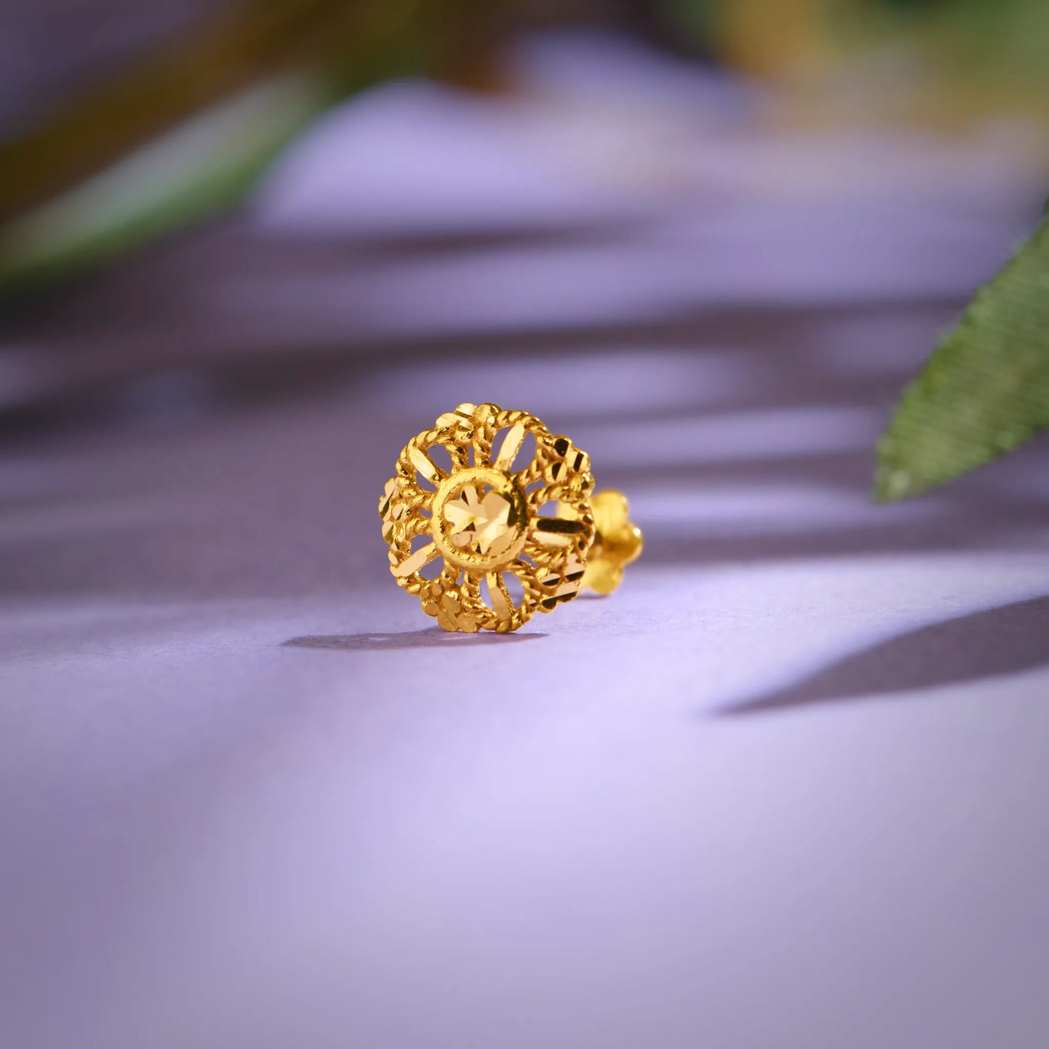 Close up of intricate floral inspired 18K gold nose pin placed on a soft lavender surface with leaf shadows highlighting fine detailing