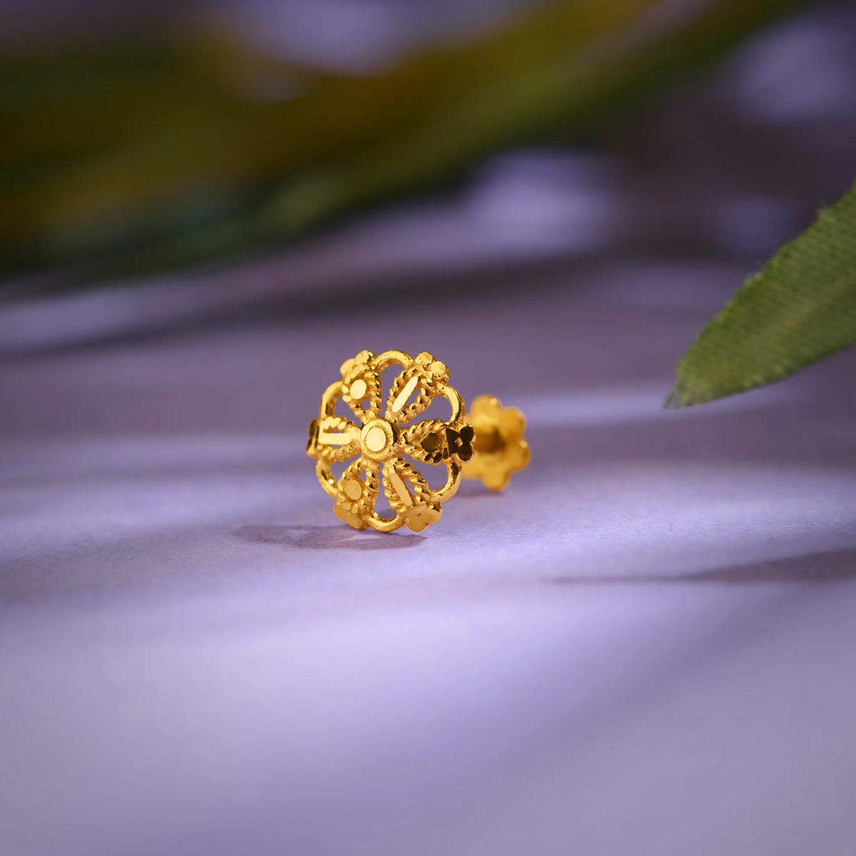 Close up of floral inspired 18K gold nose pin placed on a soft lavender surface with gentle leaf shadows highlighting intricate openwork design