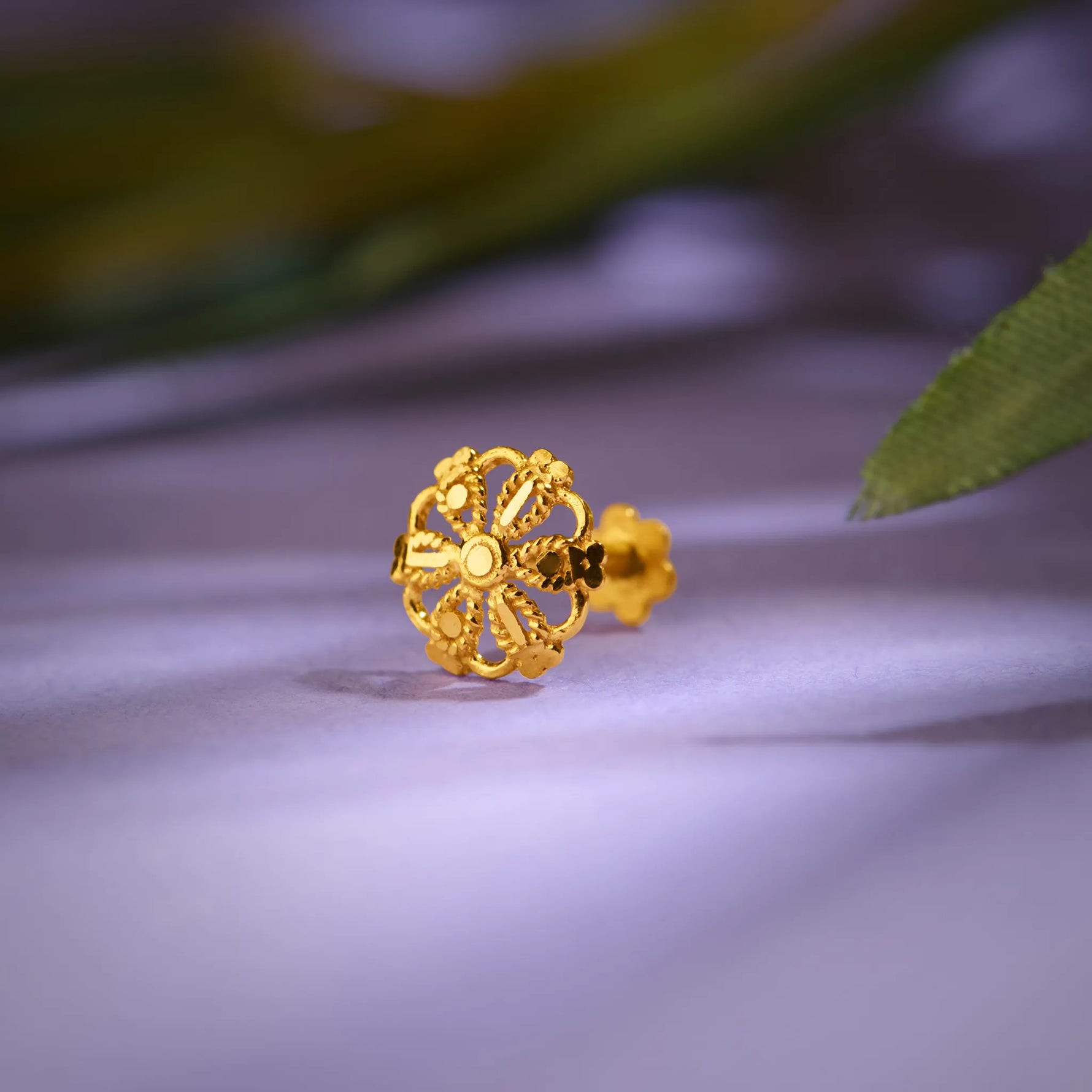Close up of floral inspired 18K gold nose pin placed on a soft lavender surface with gentle leaf shadows highlighting intricate openwork design