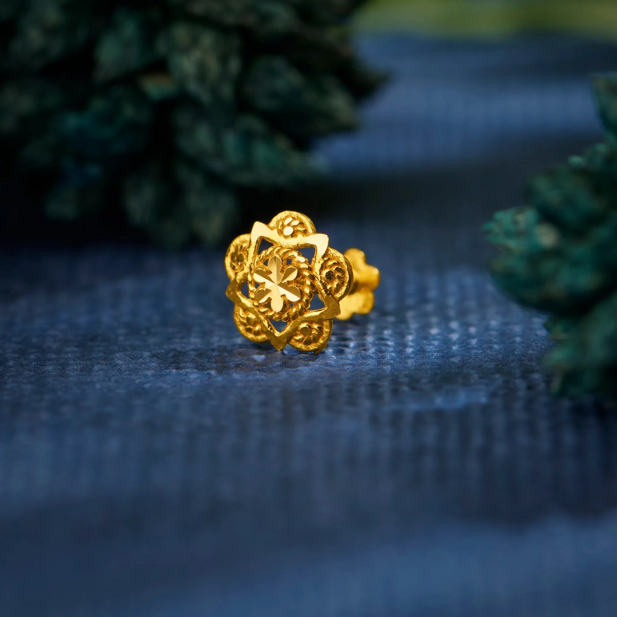 Close up view of a floral design 18K gold nose pin placed on a blue textured fabric highlighting intricate detailing