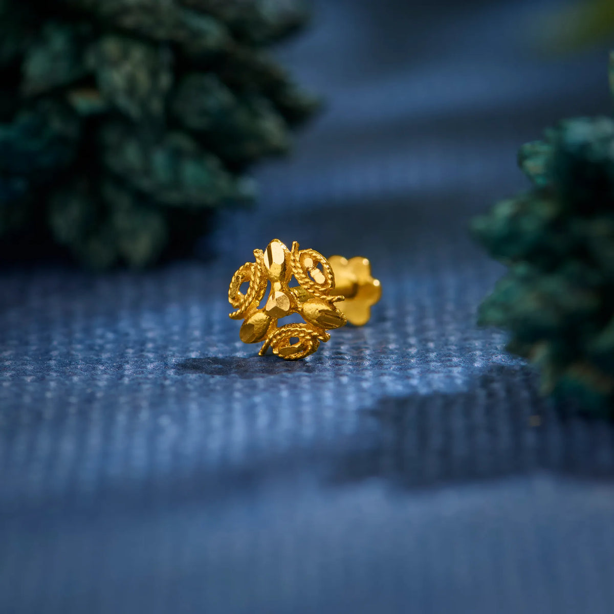 Close up view of a floral 18K gold nose pin placed on a textured blue surface highlighting intricate detailing