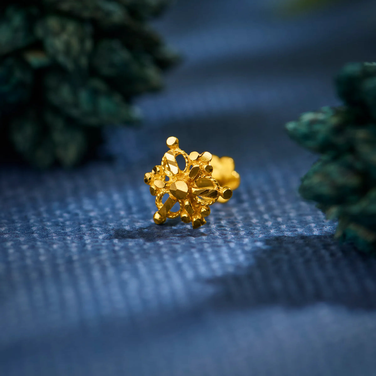 Close up of cluster style 18K gold nose pin placed on a blue textured surface showcasing its intricate floral detailing