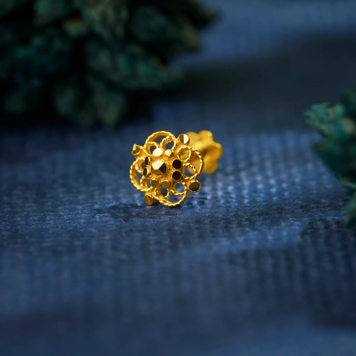 Close up of intricate floral 18K gold nose pin placed on textured blue fabric highlighting detailed craftsmanship