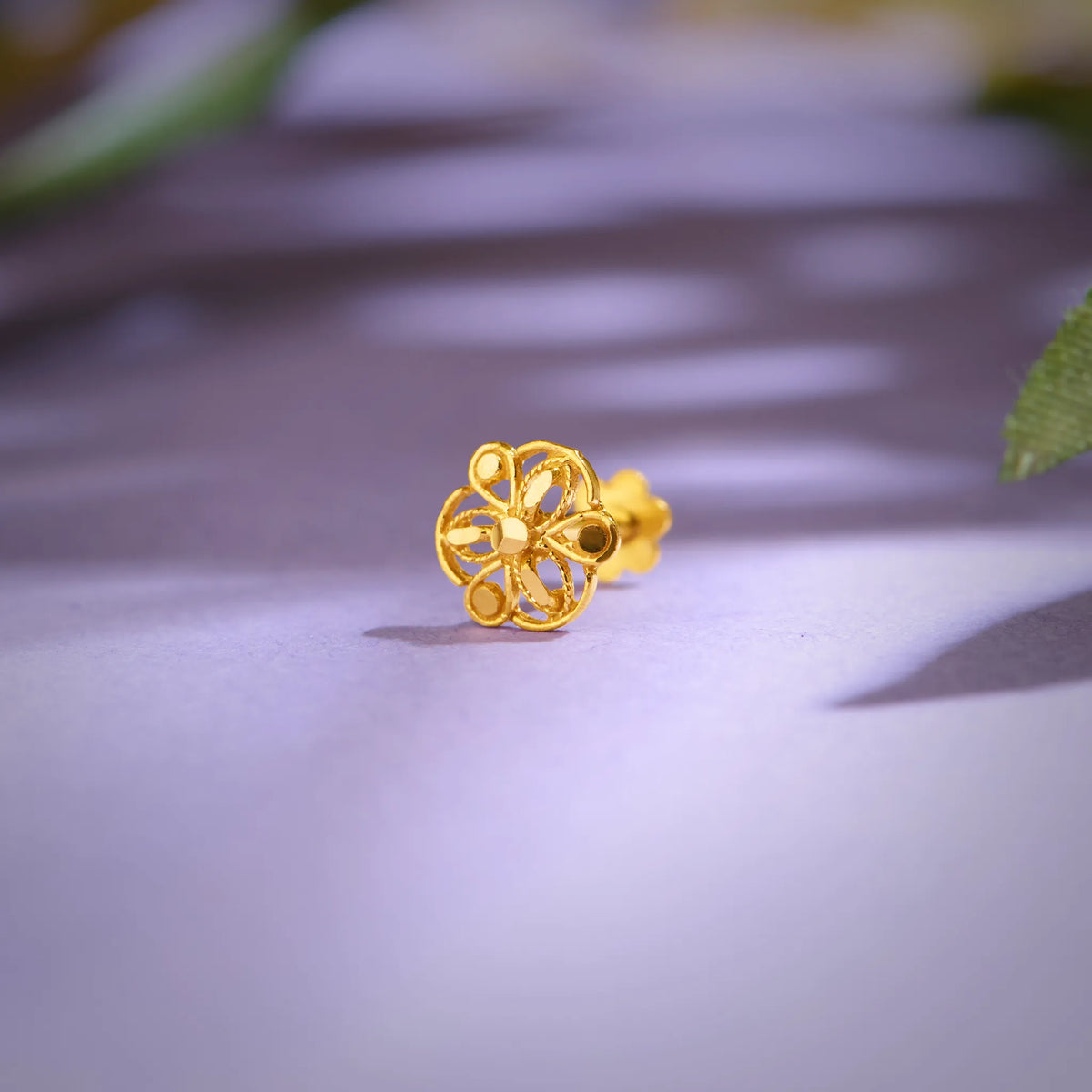 Close up of floral 18K gold nose pin placed on a soft lavender surface highlighting intricate openwork design