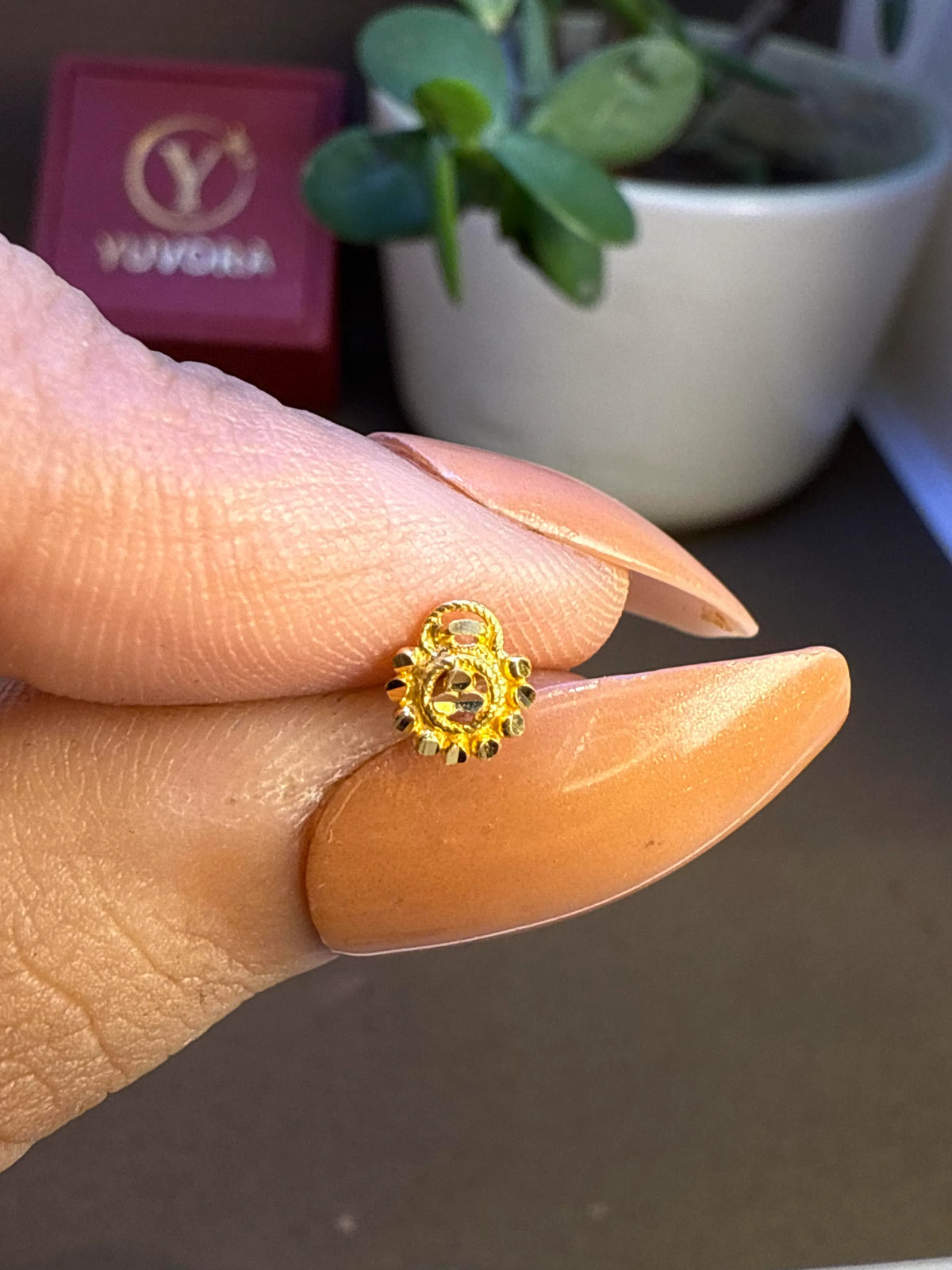 Hand holding a small floral gold nose pin near a plant pot to show its lightweight size and wearability