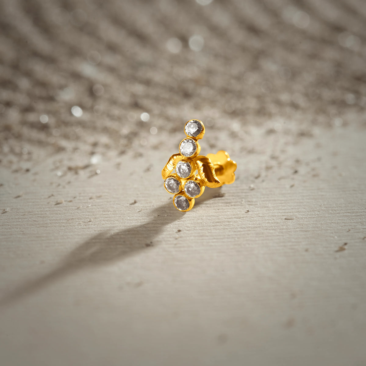 An 18K gold nose pin with an asymmetric cluster of clear crystal stones, photographed on a light textured surface