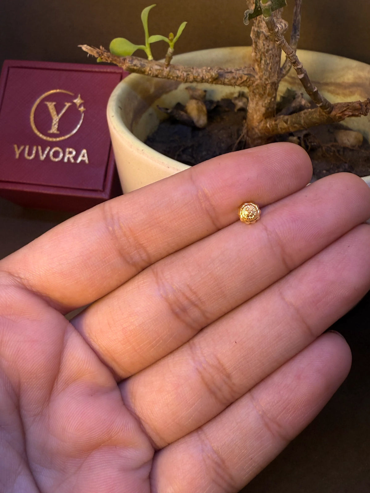 Round gold nose pin with beaded texture and crystal center, held in hand