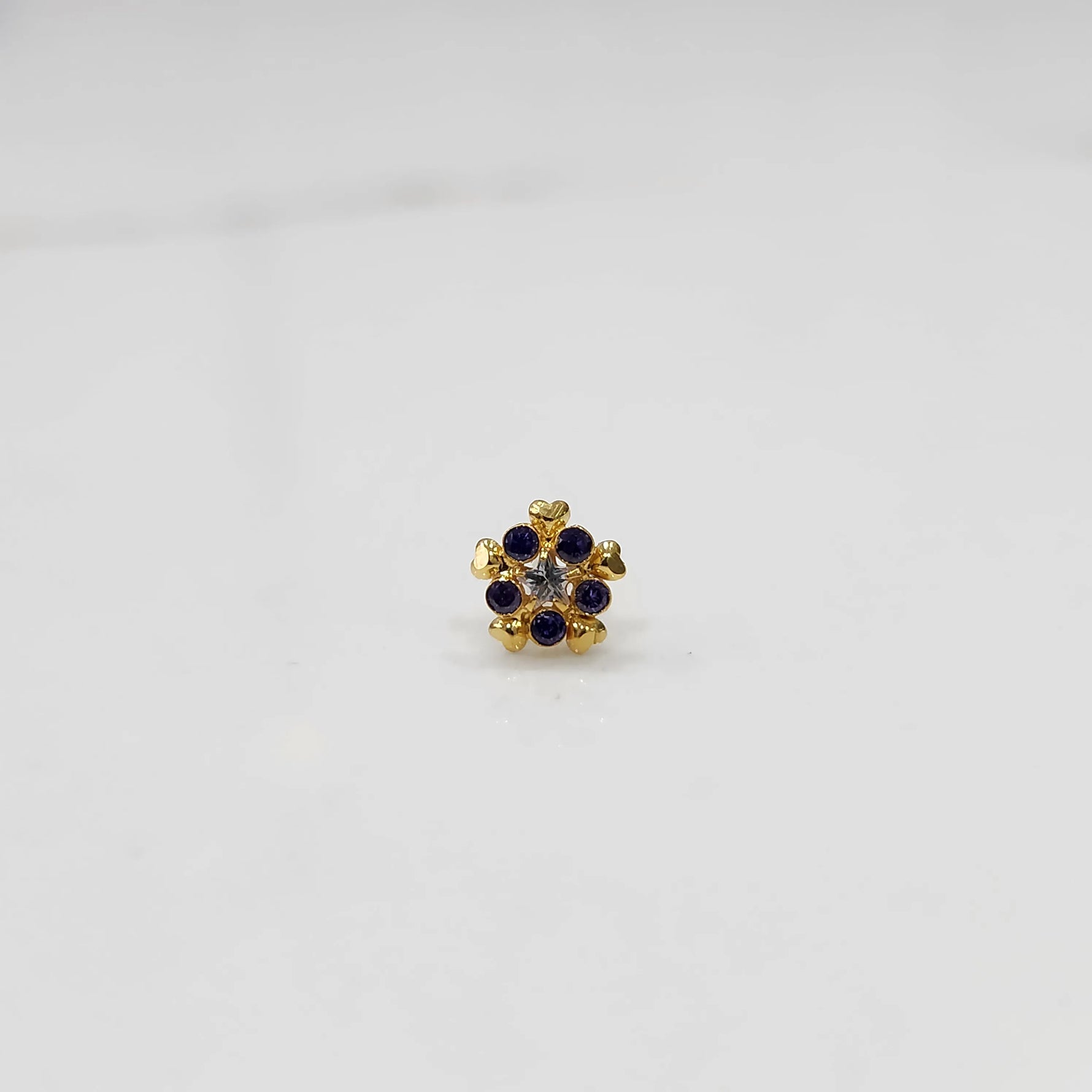 Product shot of a gold nose stud with blue sapphire gemstone flower and heart petal design