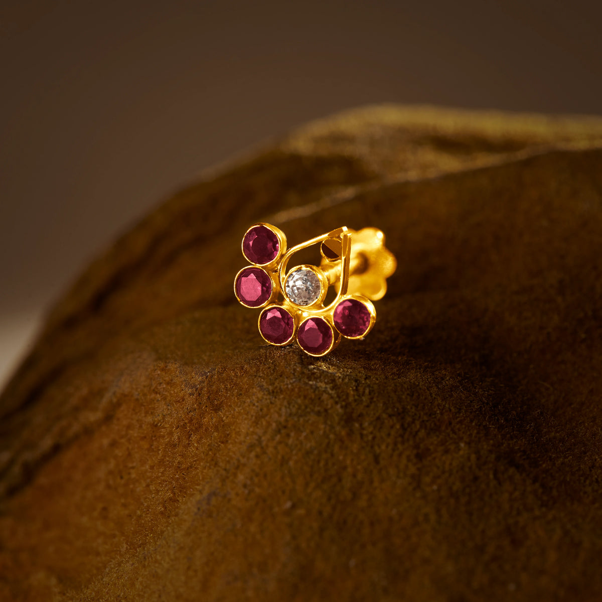 An 18K gold nose pin shaped like a fan, set with ruby-red stones and a single clear crystal center, photographed on a dark textured fabric background