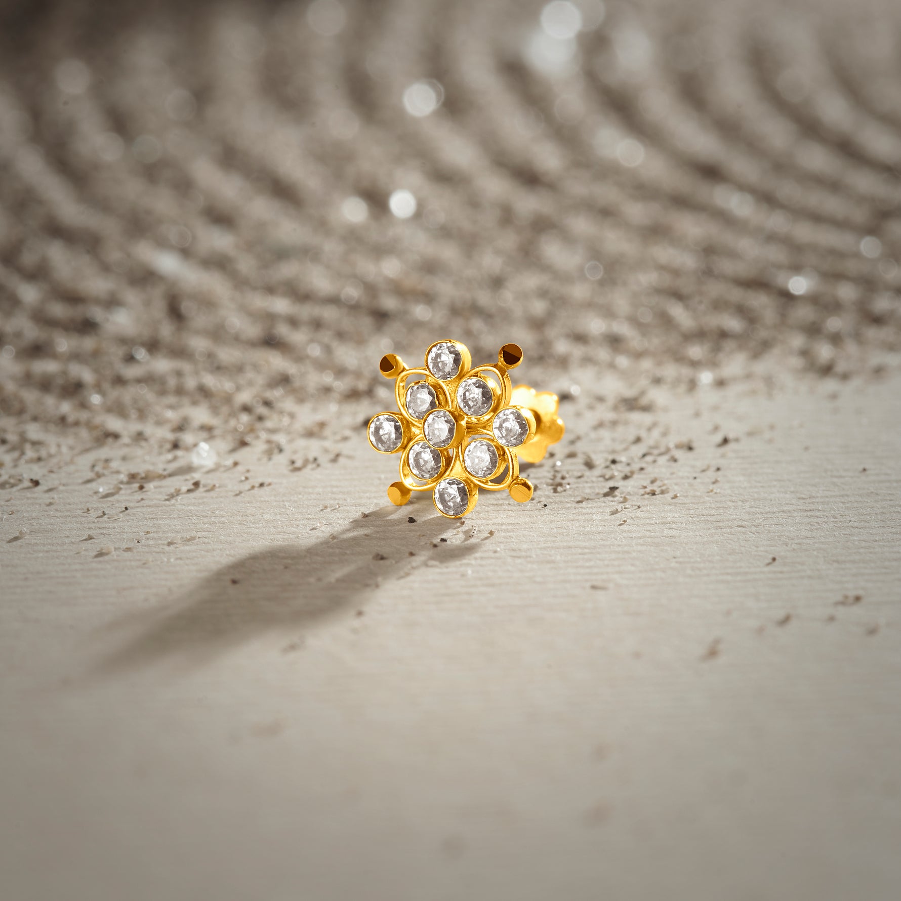 A gold nose pin shaped like a flower, set with clear crystal stones, styled on a textured surface