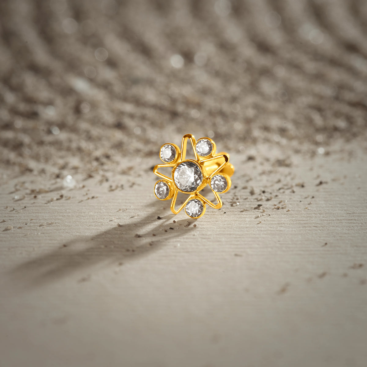 A gold nose pin shaped like a star, set entirely with clear crystal stones, photographed on a textured sandy surface