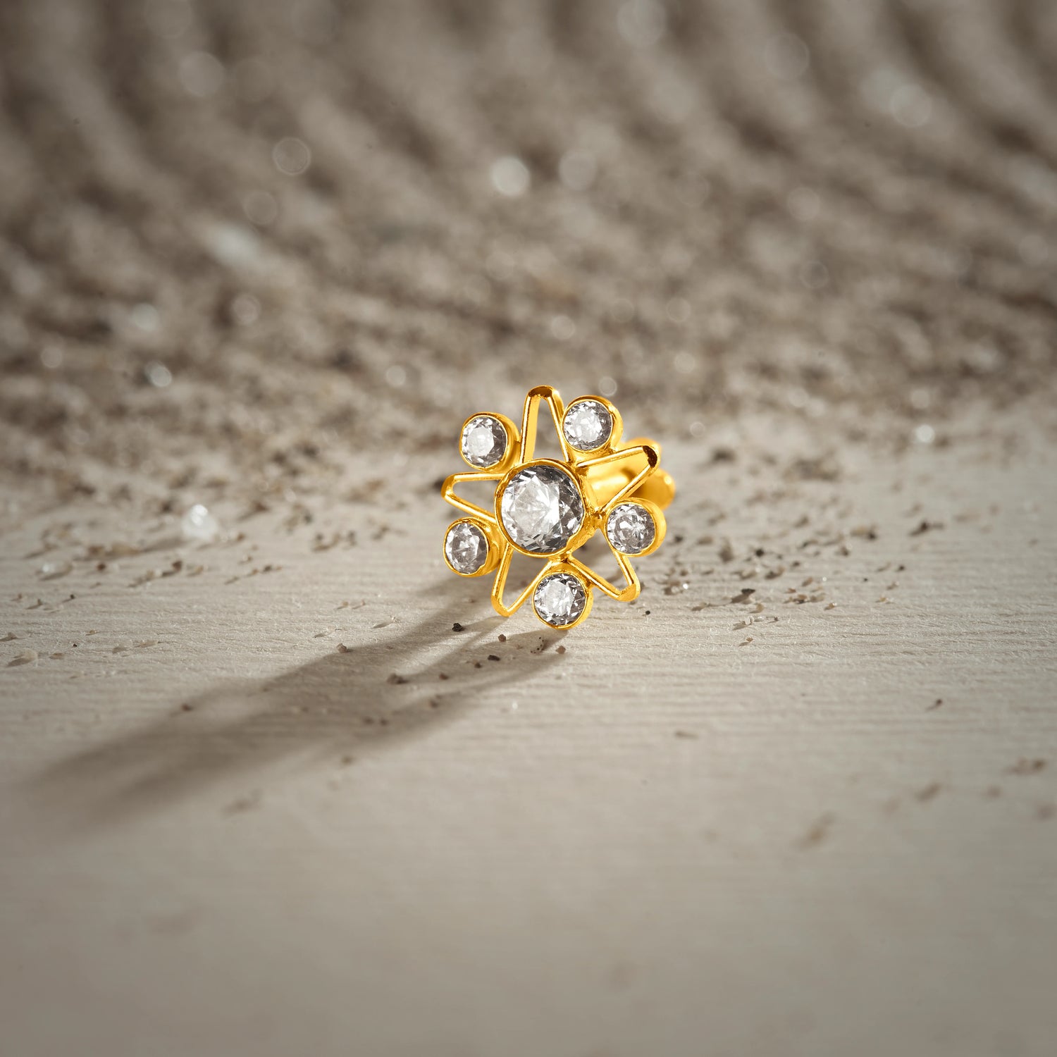 A gold nose pin shaped like a star, set entirely with clear crystal stones, photographed on a textured sandy surface