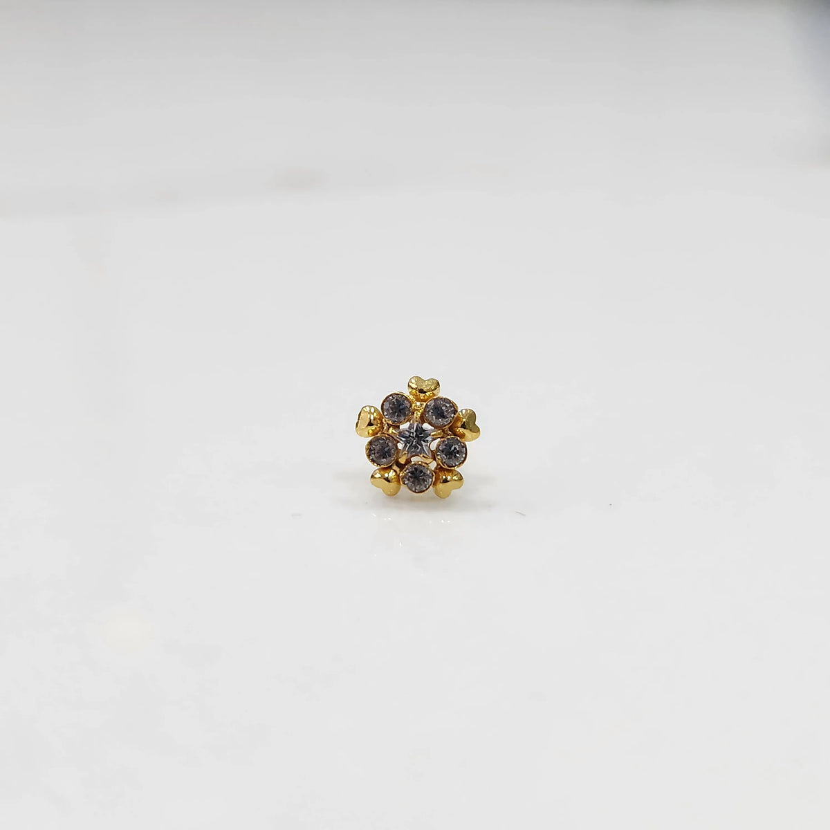 Product shot of a gold nose stud with clear gemstone flower and heart petal design