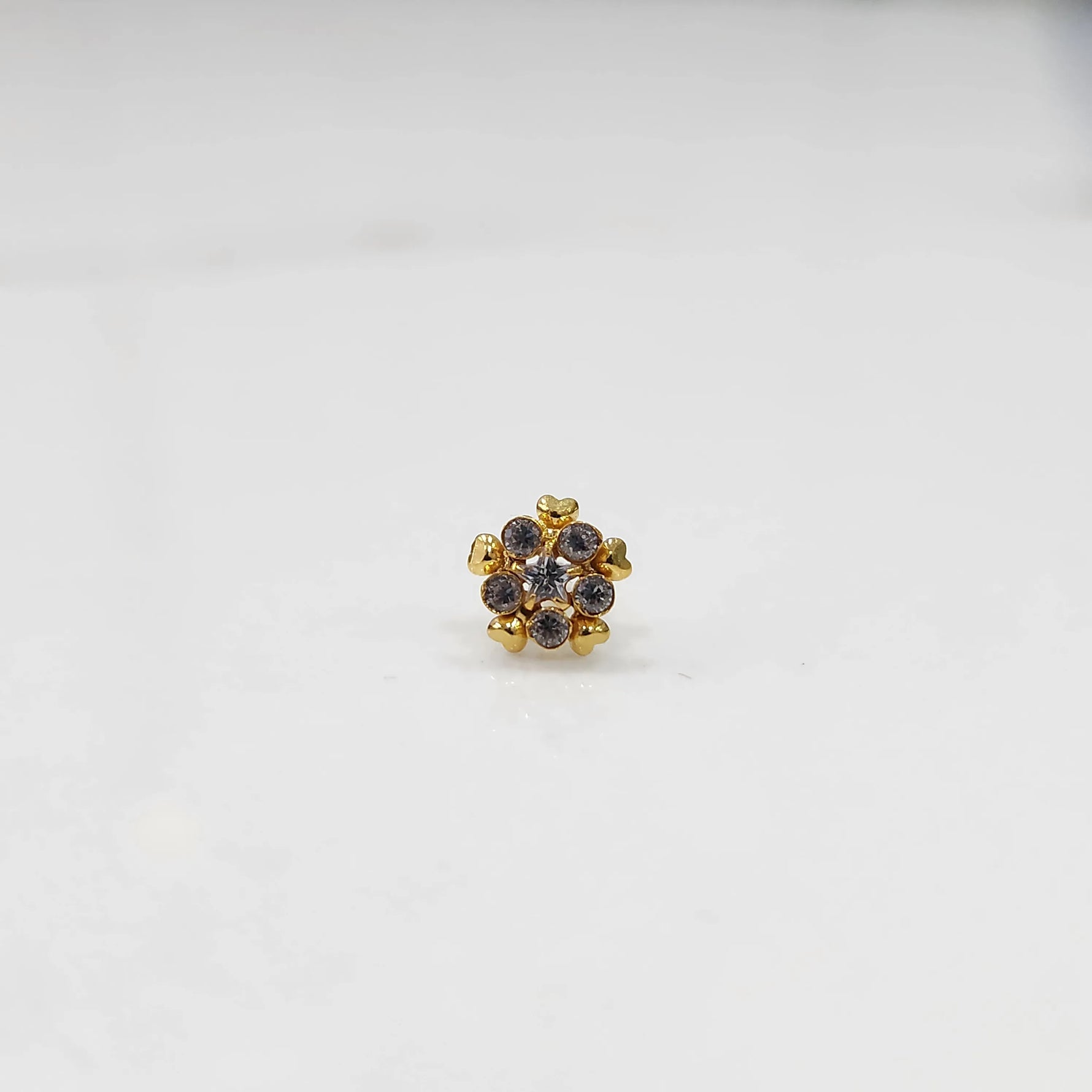 Product shot of a gold nose stud with clear gemstone flower and heart petal design