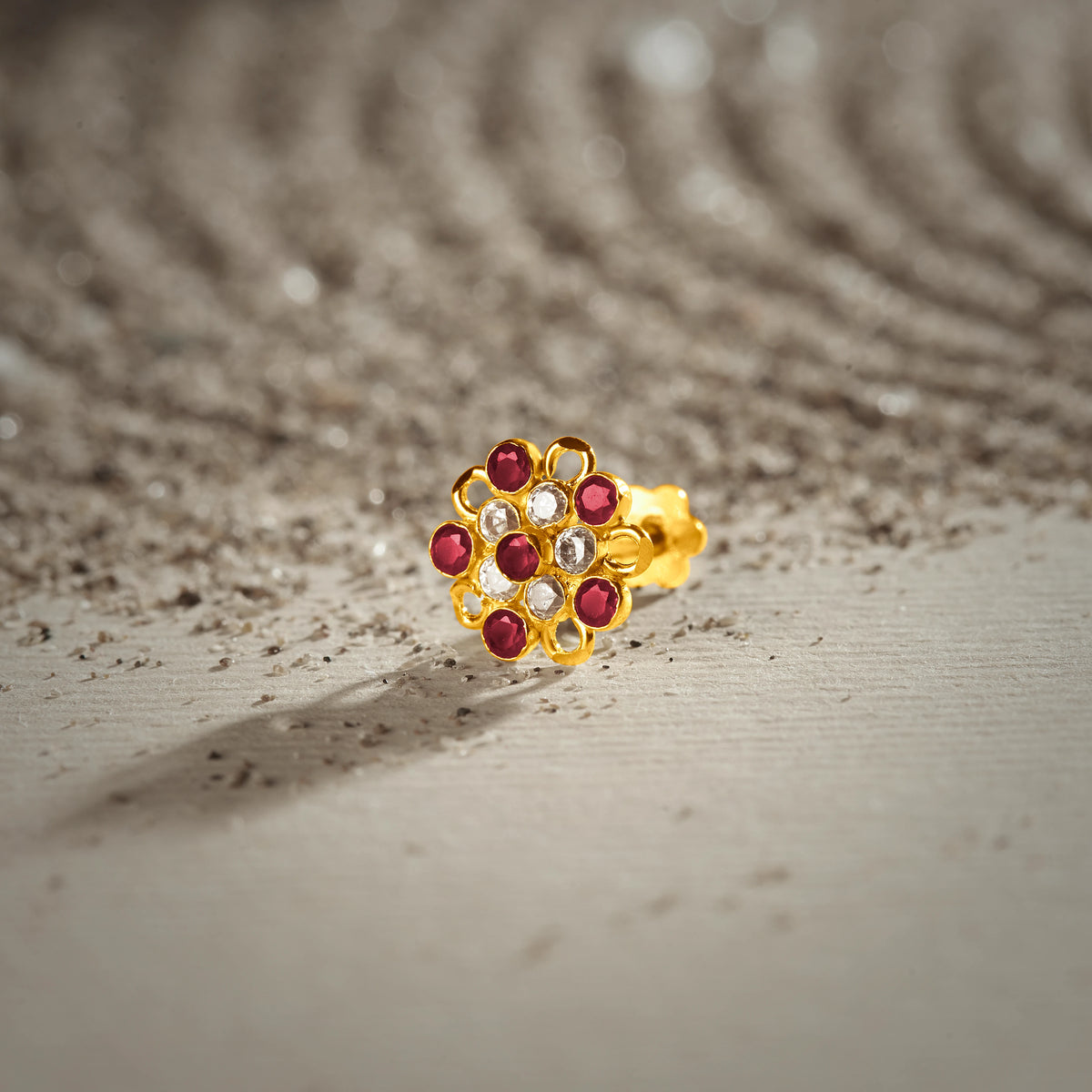 An 18K gold nose pin shaped like a daisy flower with open petal outlines, set with alternating ruby-red and clear crystal stones, photographed on a light textured surface