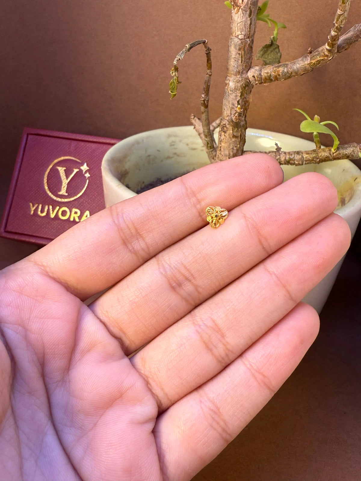 Round flower-shaped gold nose pin with center stone held in hand, Yuvora jewelry packaging in background