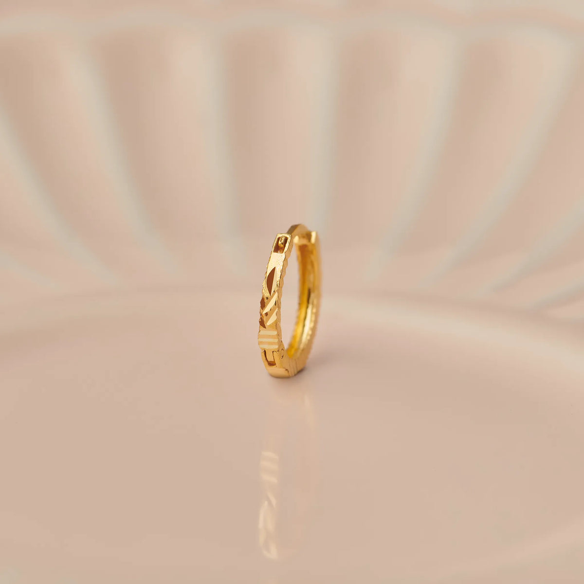 Side angle view of a slim textured 18K gold hoop nose ring placed on a soft beige surface with gentle curved light patterns